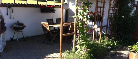 Un patio con mesa y sillas de madera, una parrilla y un jardín con plantas trepadoras y flores.