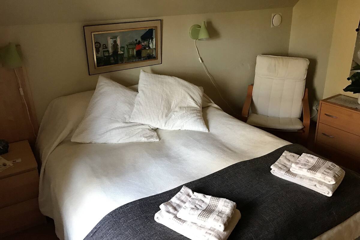 Ein ordentlich bezogenes Bett mit weißen Leinen, ein Sessel, ein Nachttisch und ein Gemälde an der Wand.