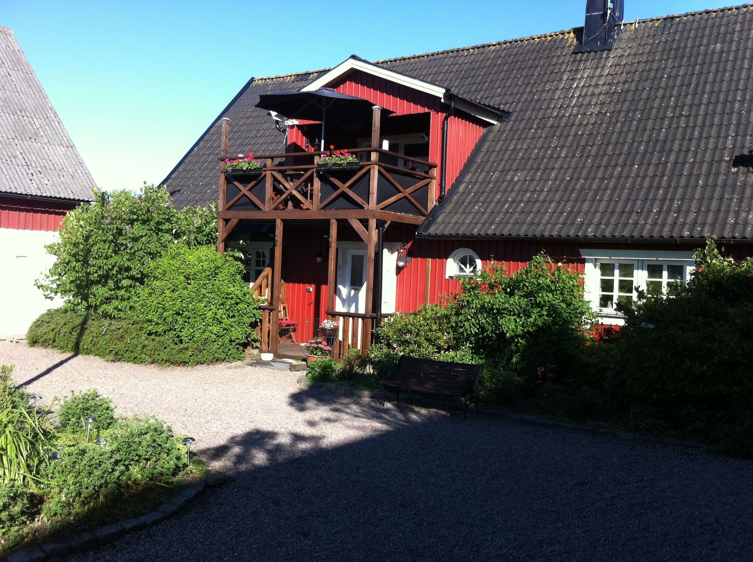 A red building with a balcony, gravel driveway, and surrounding greenery.