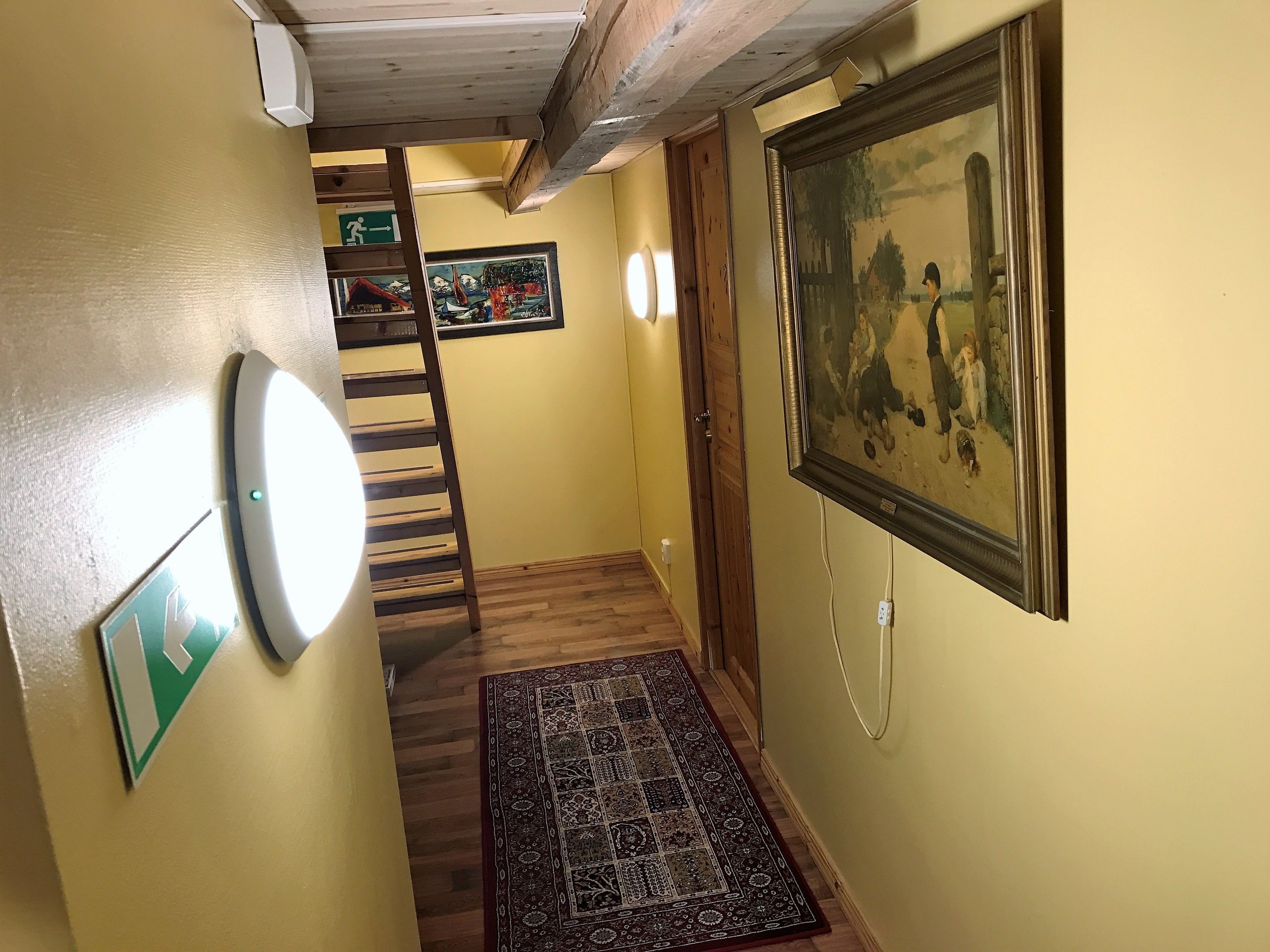 Un couloir avec un escalier en bois, un tapis à motifs, un tableau encadré et un luminaire fixé au mur.