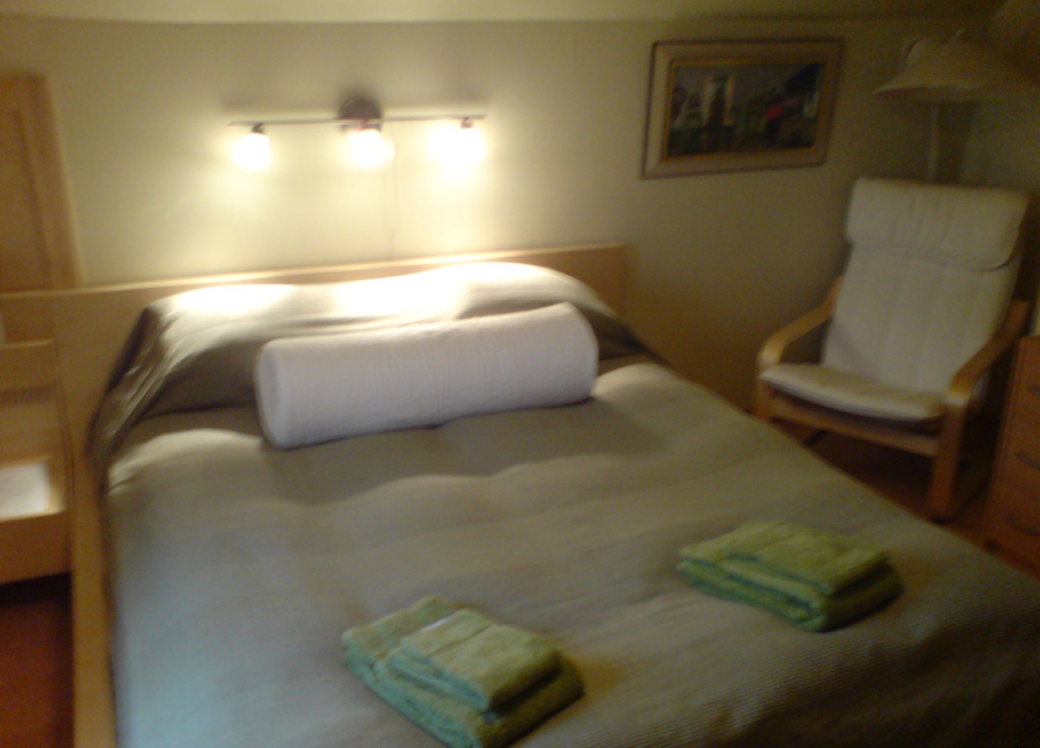 Una cama bien hecha con una almohada y dos toallas dobladas, una silla de madera y una lámpara fijada a la pared.