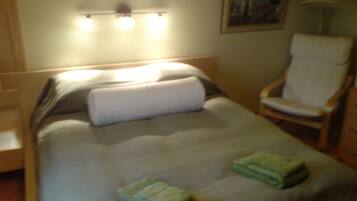 Una cama bien hecha con una almohada y dos toallas dobladas, una silla de madera y una lámpara fijada a la pared.