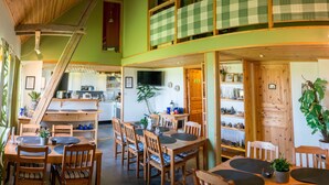 A dining area with wooden tables and chairs, a kitchen in the background, and a checkered balcony. - Mattisgården Bed & Breakfast (Klippan)