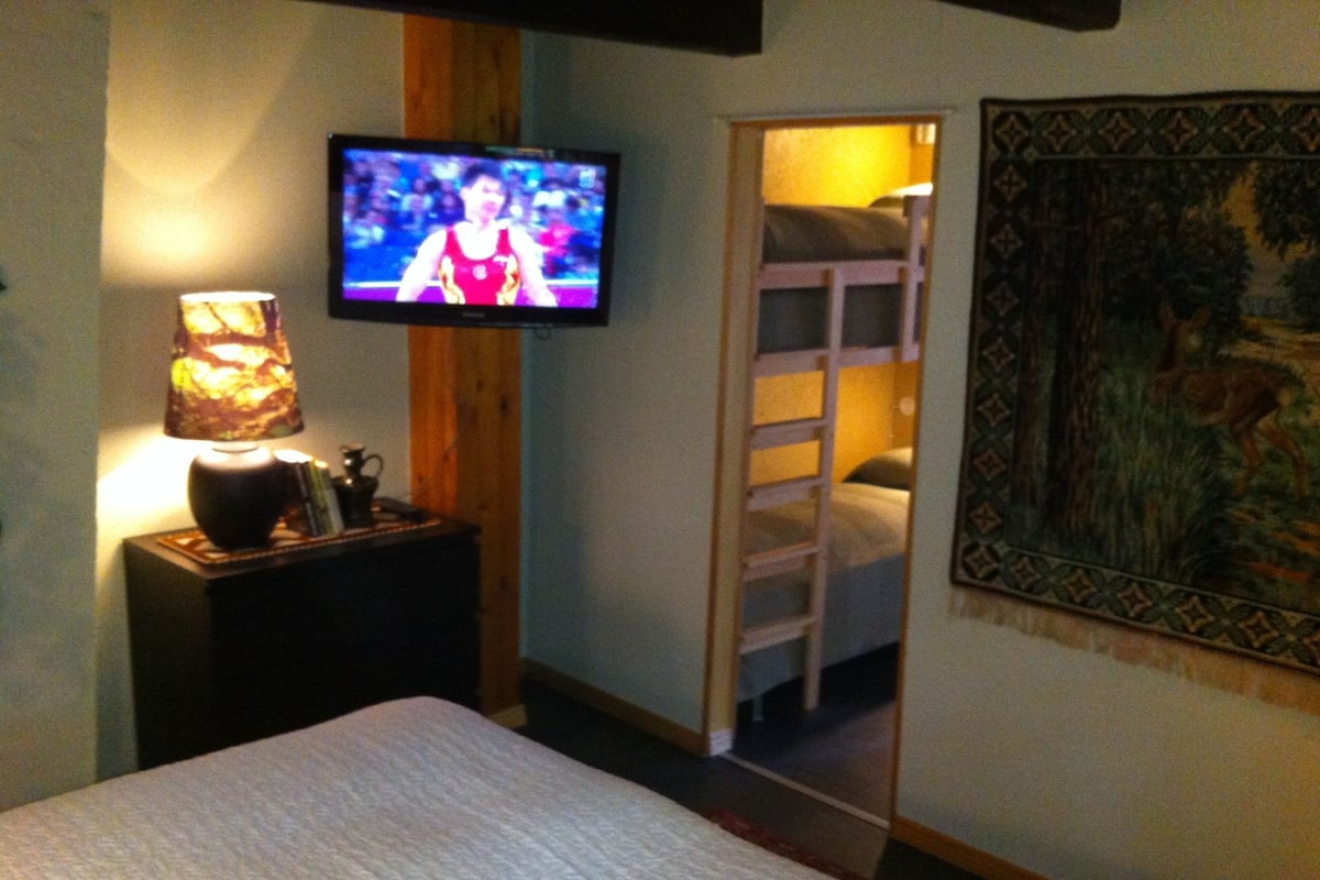 Ein Schlafzimmer mit einem Bett, einem an der Wand montierten Fernseher, einem Nachttisch mit Lampe, einem Etagenbett und einem Wandteppich.