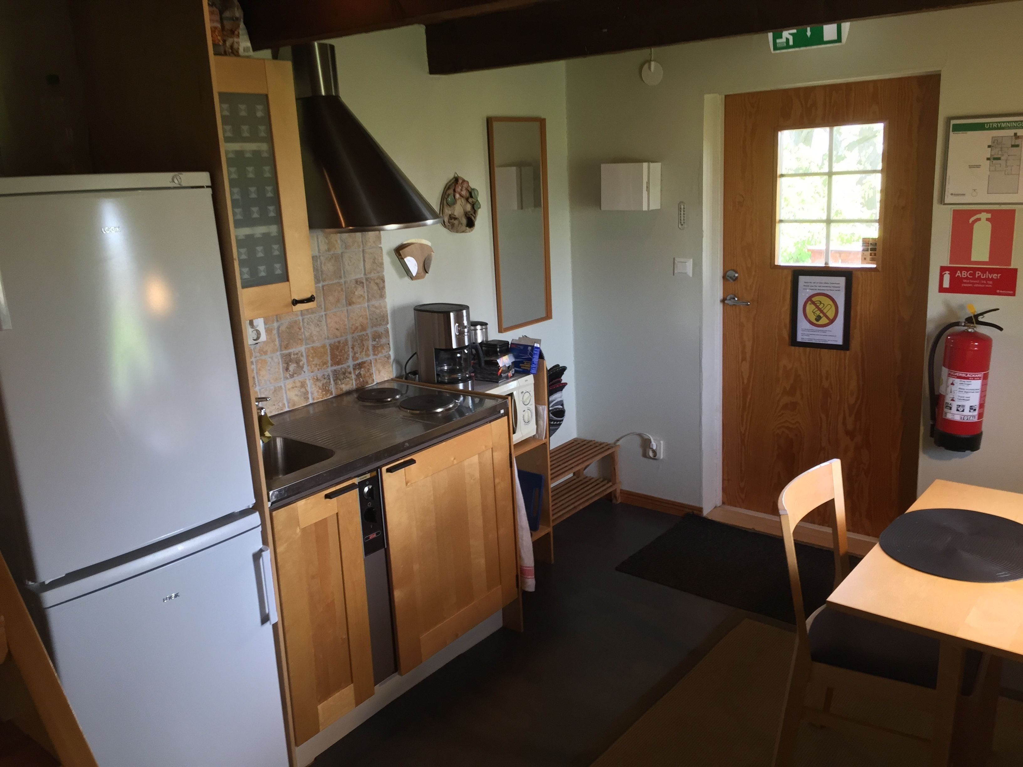 A compact kitchen with a refrigerator, stove, and sink. There is a wooden cabinet, a mirror, and a fire extinguisher on the wall.