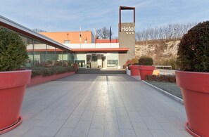Entrada d'un hotel modern amb façana de vidre, un rètol destacat i un camí pavimentat amb teules vermelles.