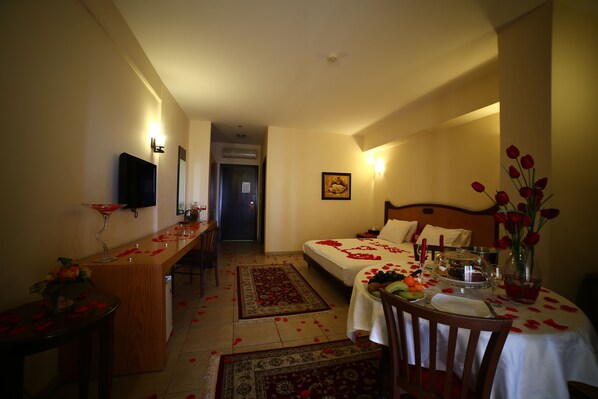 Habitación de hotel con cama, escritorio, silla, mesa de comedor y un jarrón de flores, además de un televisor en la pared.
