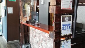 Ein Empfangstresen mit Holzdetaillierung, einer Glastrennwand und verschiedenen Hinweisschildern, darunter Maßnahmen zur COVID-19-Vorbeugung.