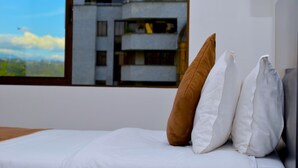 Ein Schlafzimmer mit einem Bett, weißer Bettwäsche und zwei Kissen. Ein Fenster gibt den Blick frei auf ein modernes Gebäude mit Balkonen.