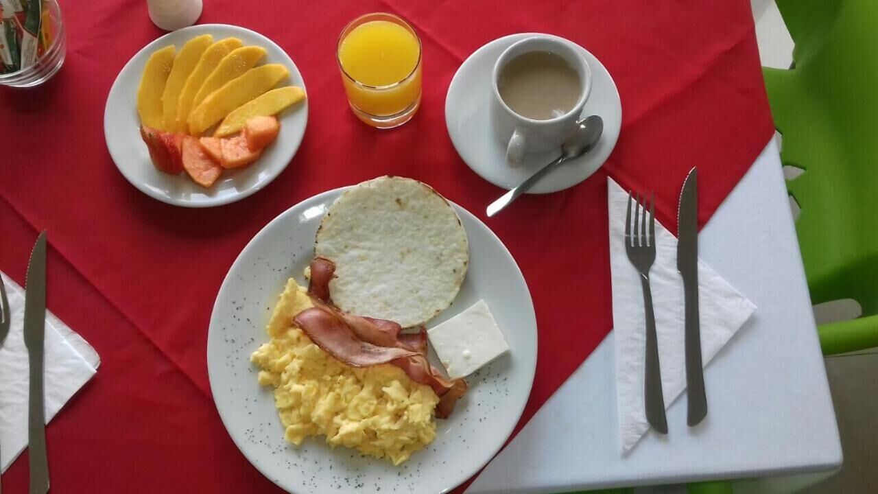 A breakfast setting with a plate of scrambled eggs, bacon, and cheese, a plate of sliced fruit, a glass of orange juice, and a cup of coffee on a red tablecloth.