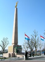 Un monumento alto con una estatua en la parte superior, rodeado de árboles desprovistos de hojas y dos banderas.
