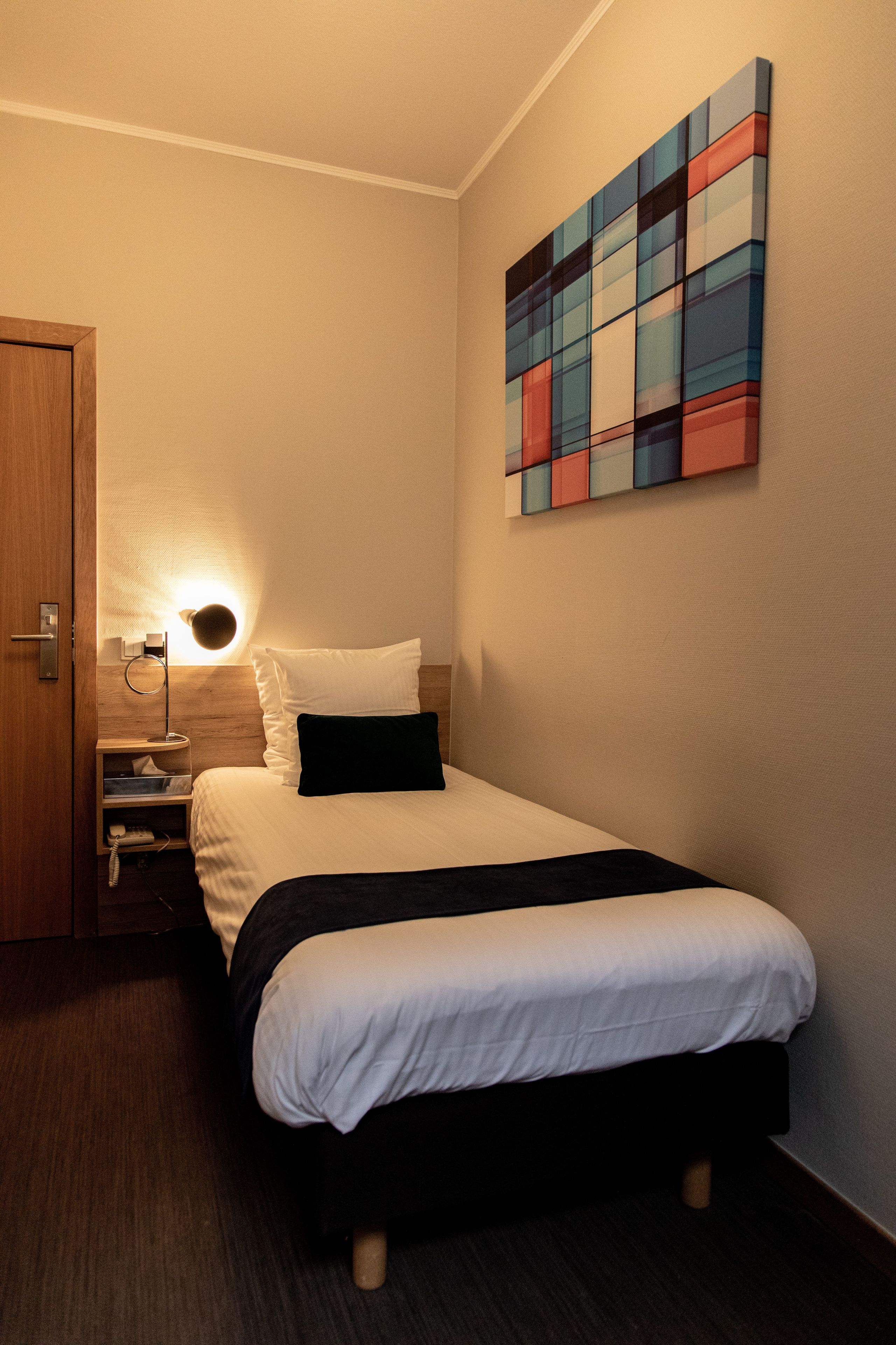 A hotel room with a bed, a bedside lamp, a nightstand, and a wall-mounted artwork.