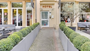 Entrada de hotel con un camino pavimentado bordeado por setos cuidados, una puerta de vidrio transparente y un cartel con el nombre del hotel.