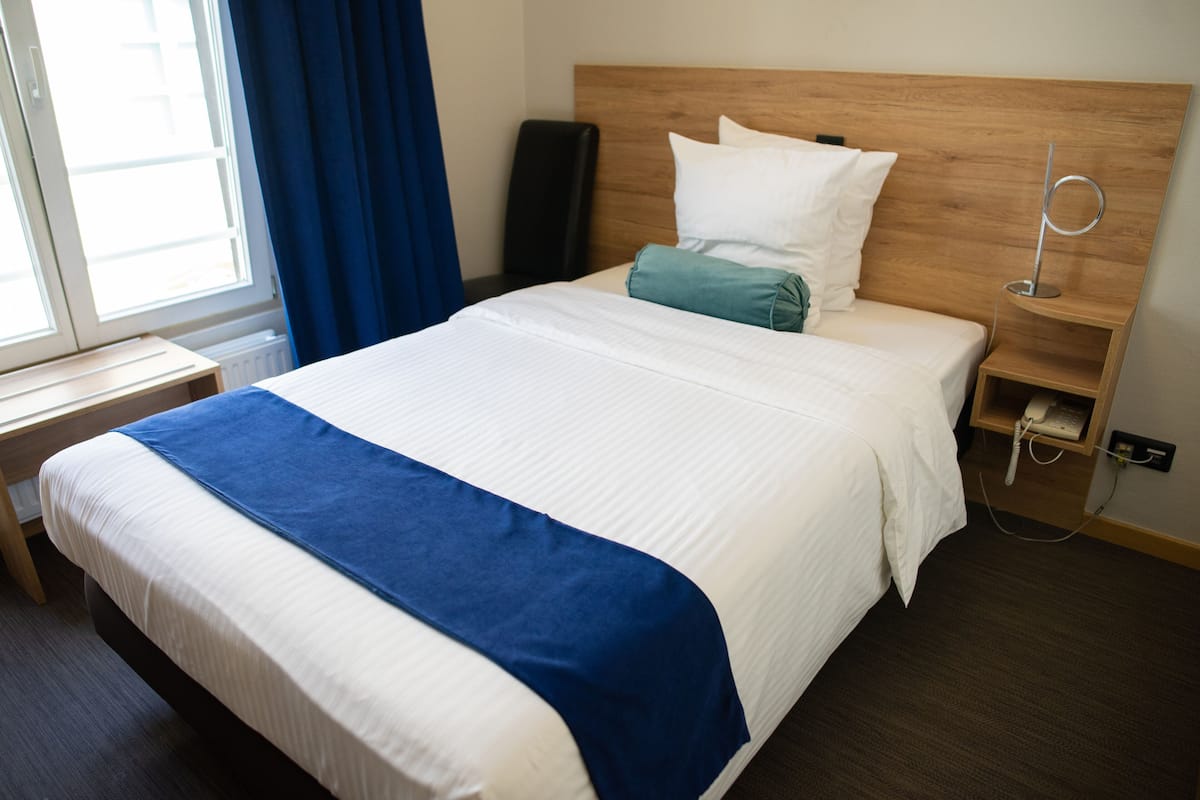 Ein ordentlich bezogenes Bett mit blauem Akzent, ein hölzernes Kopfende, ein Nachttisch mit Telefon und ein Fenster mit Vorhängen.