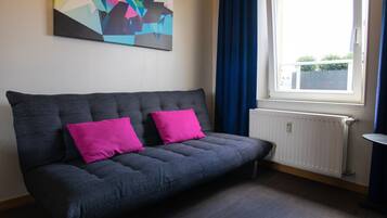 A grey sofa with two bright pink cushions, a colorful abstract painting on the wall, a window with blue curtains, and a white radiator.