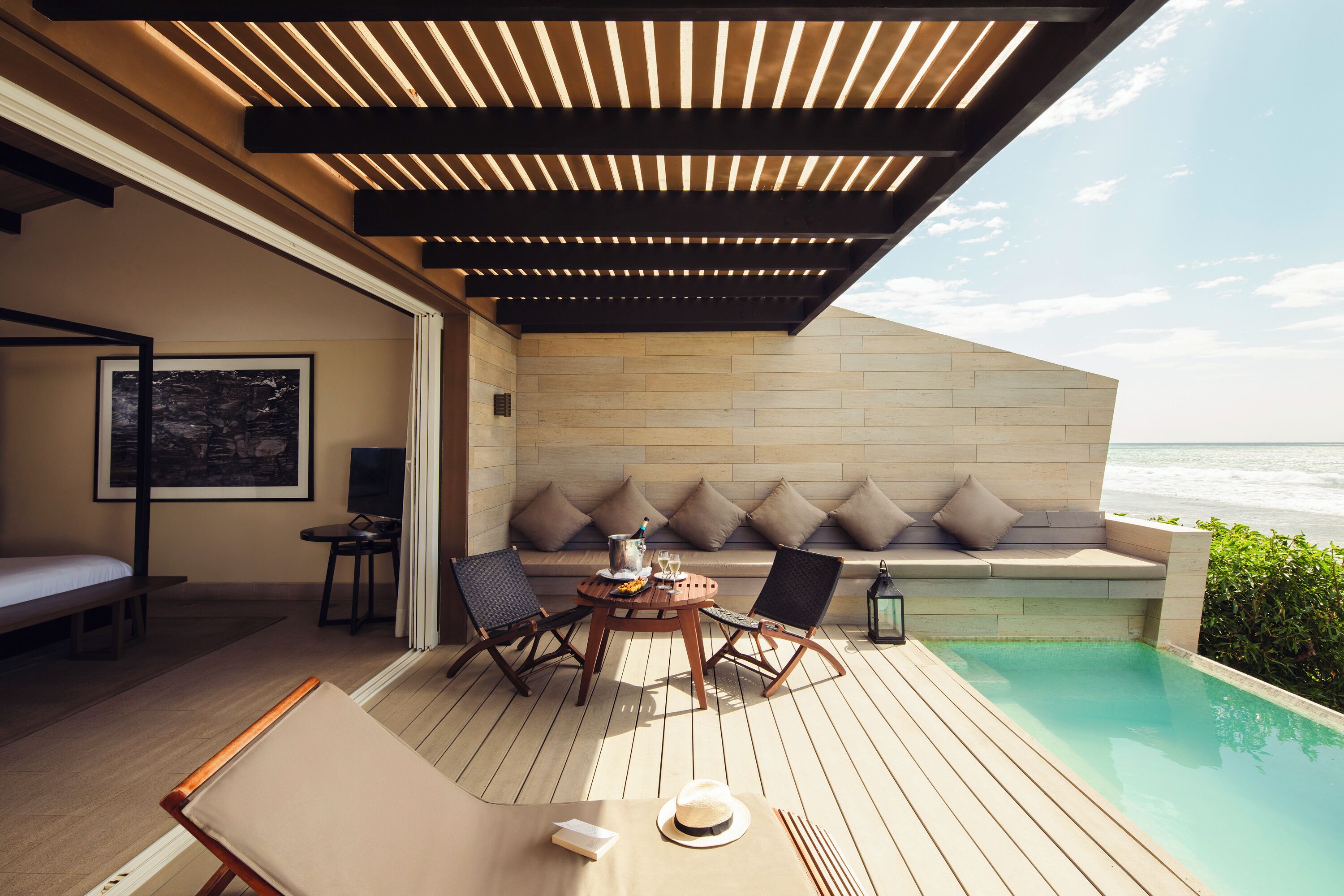 A wooden deck with a pool, a seating area with cushions, and a view of the ocean.
