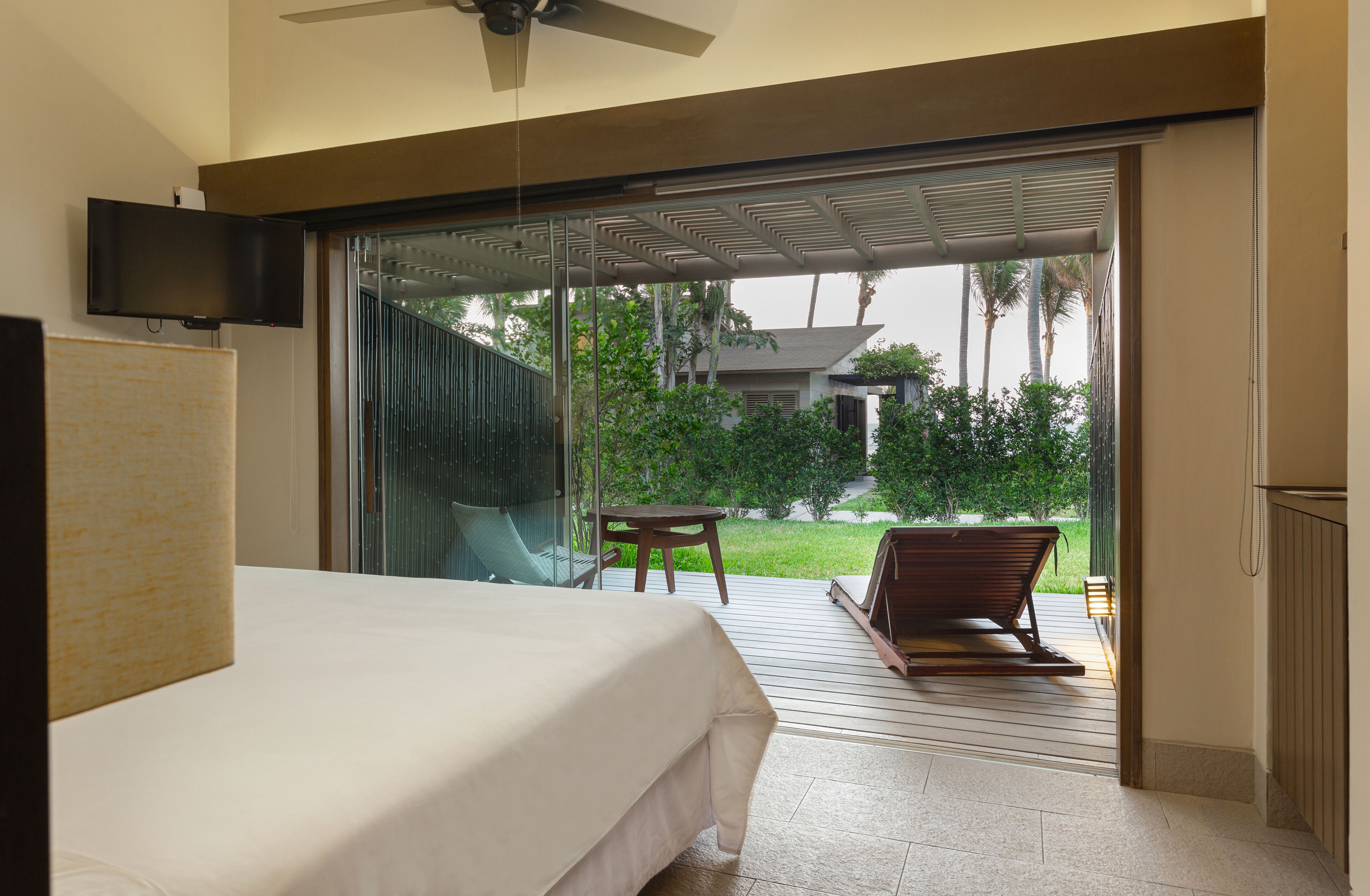 A hotel room with a bed, a television mounted on the wall, and a sliding glass door leading to an outdoor area with a table and a bench.