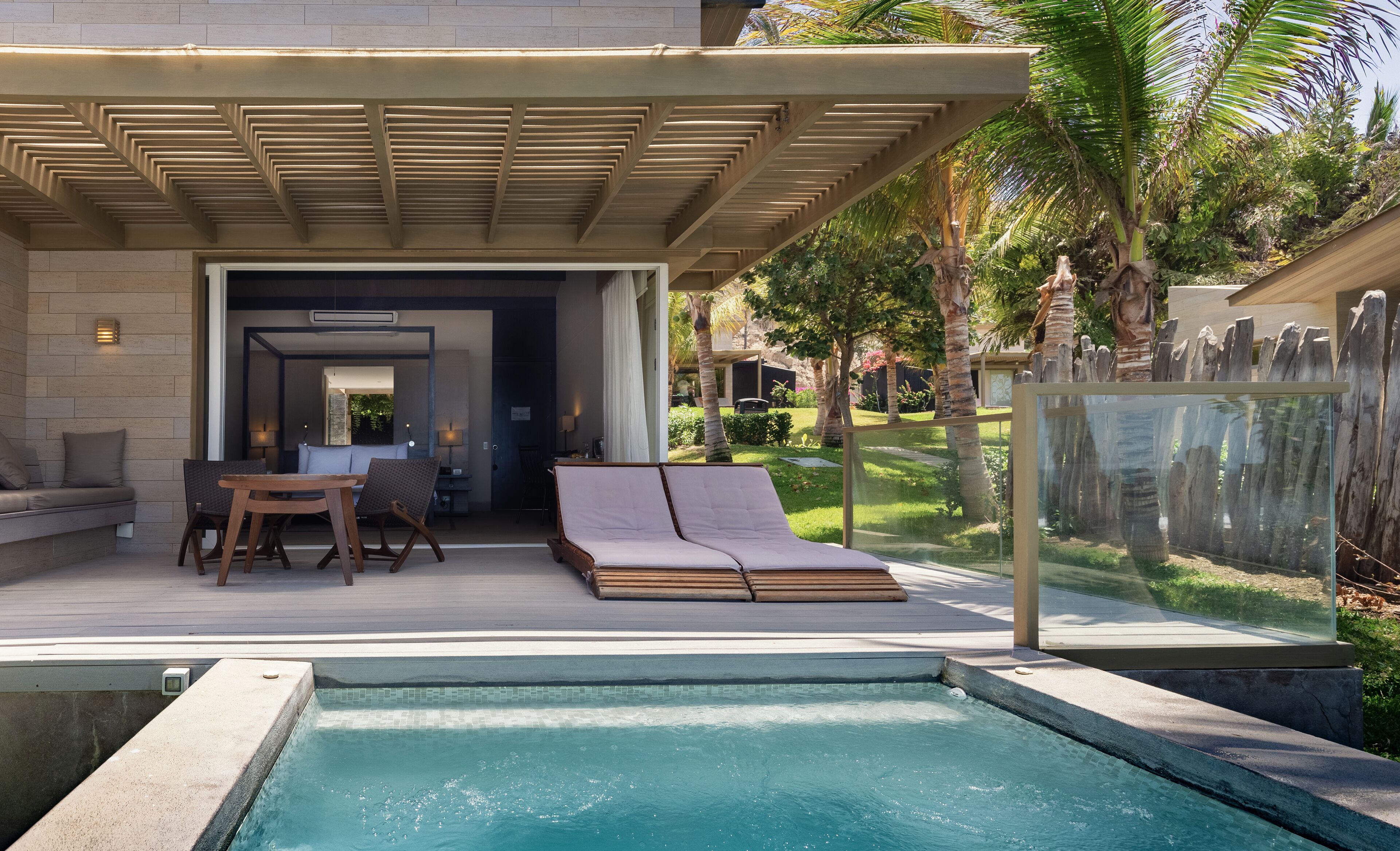 Un área de piscina moderna al aire libre con una zona de estar cubierta, una silla de descanso y vistas a un jardín exuberante.