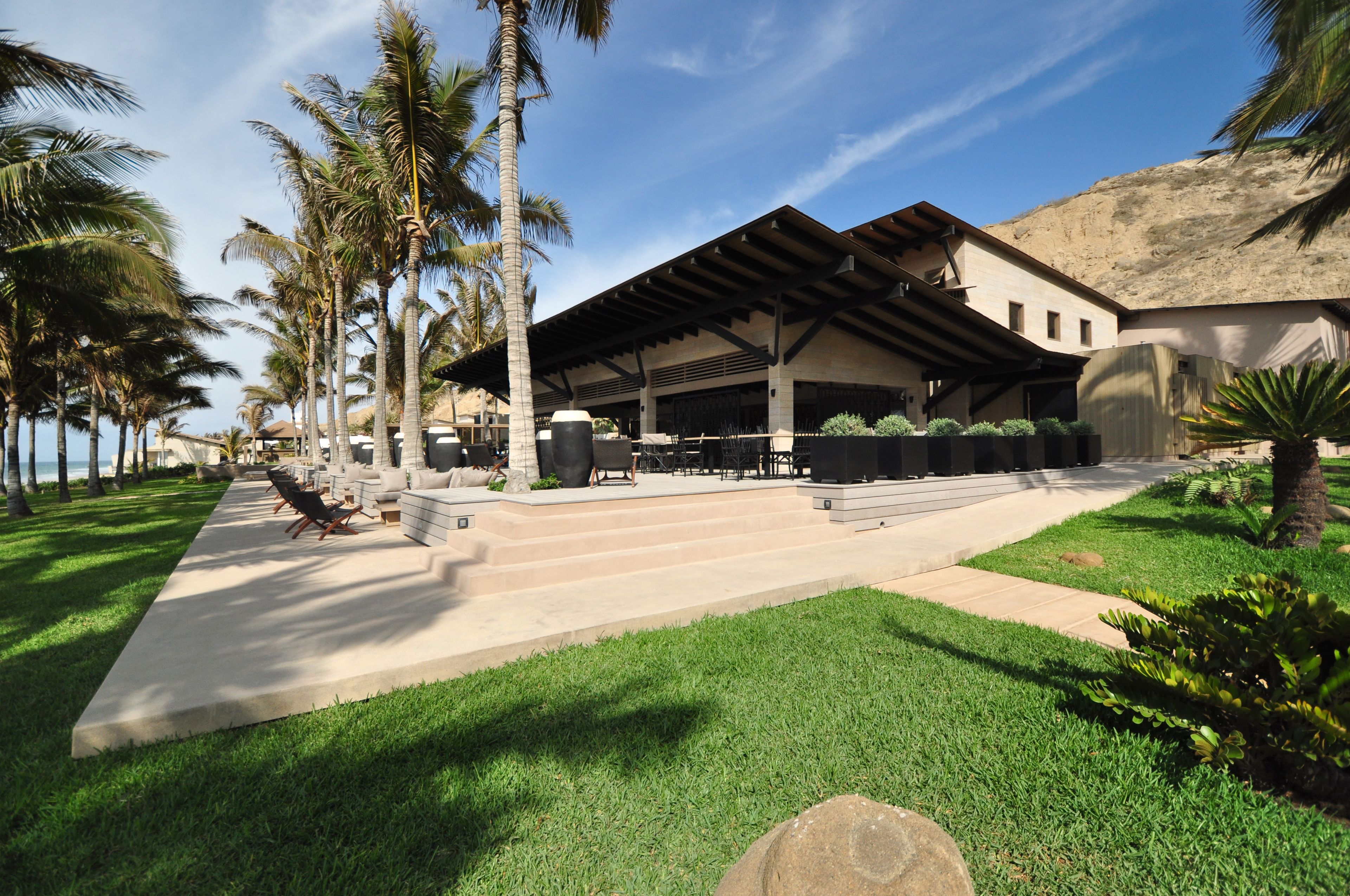 A resort with a covered outdoor seating area, palm trees, and a clear sky.