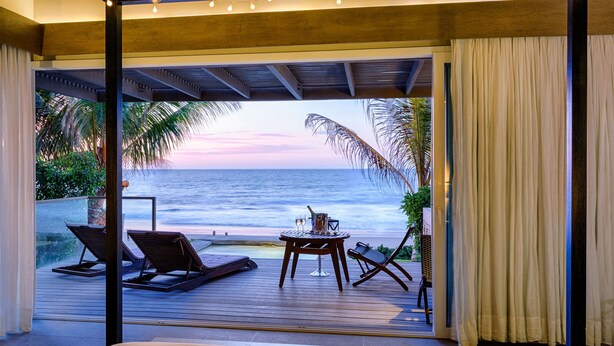 A bedroom with a large window overlooking a beach at sunset.