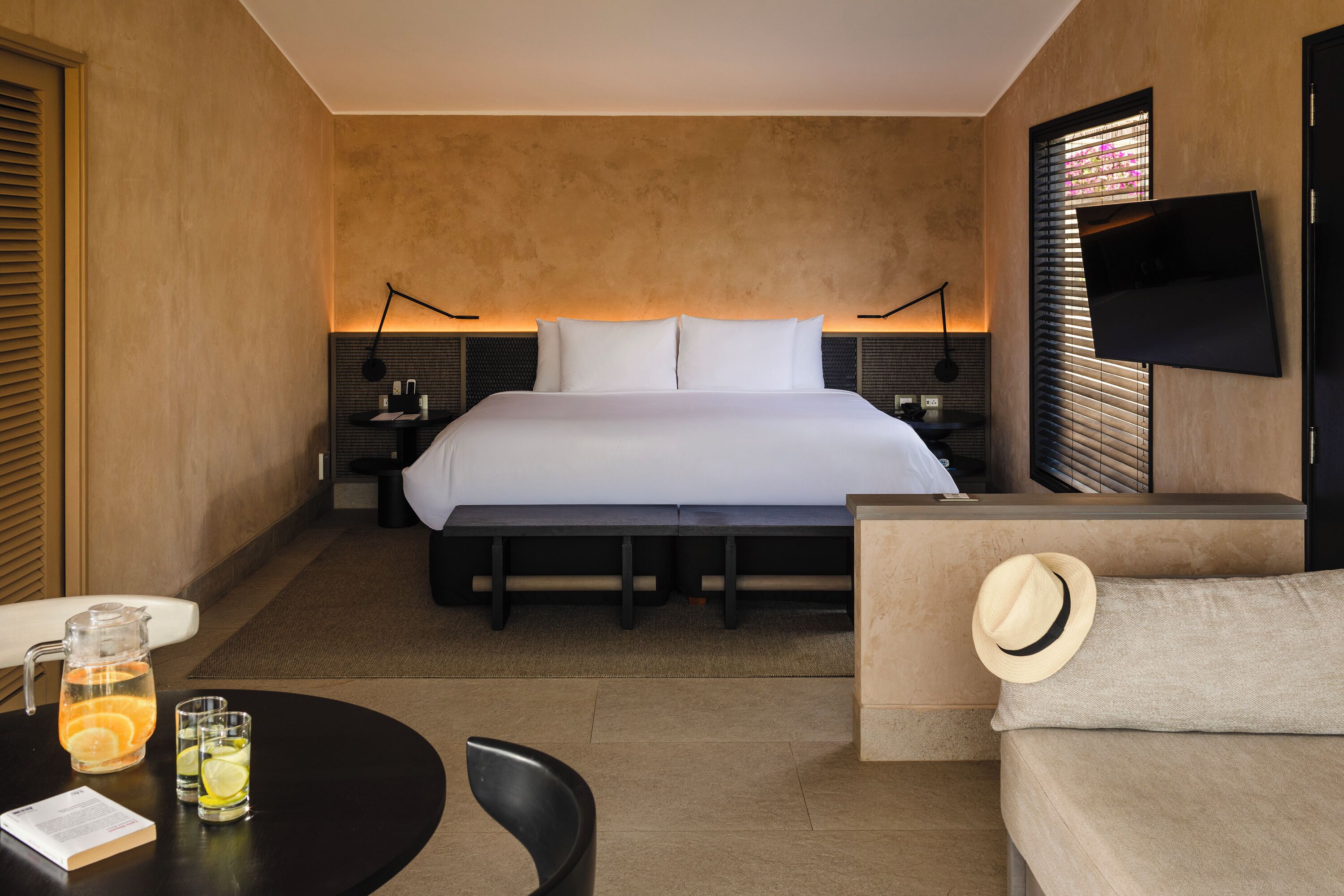 Habitación de hotel con una cama grande, mesitas de noche, televisión, zona de estar con sofá y sillón, y una pequeña mesa con jarrón y vasos.