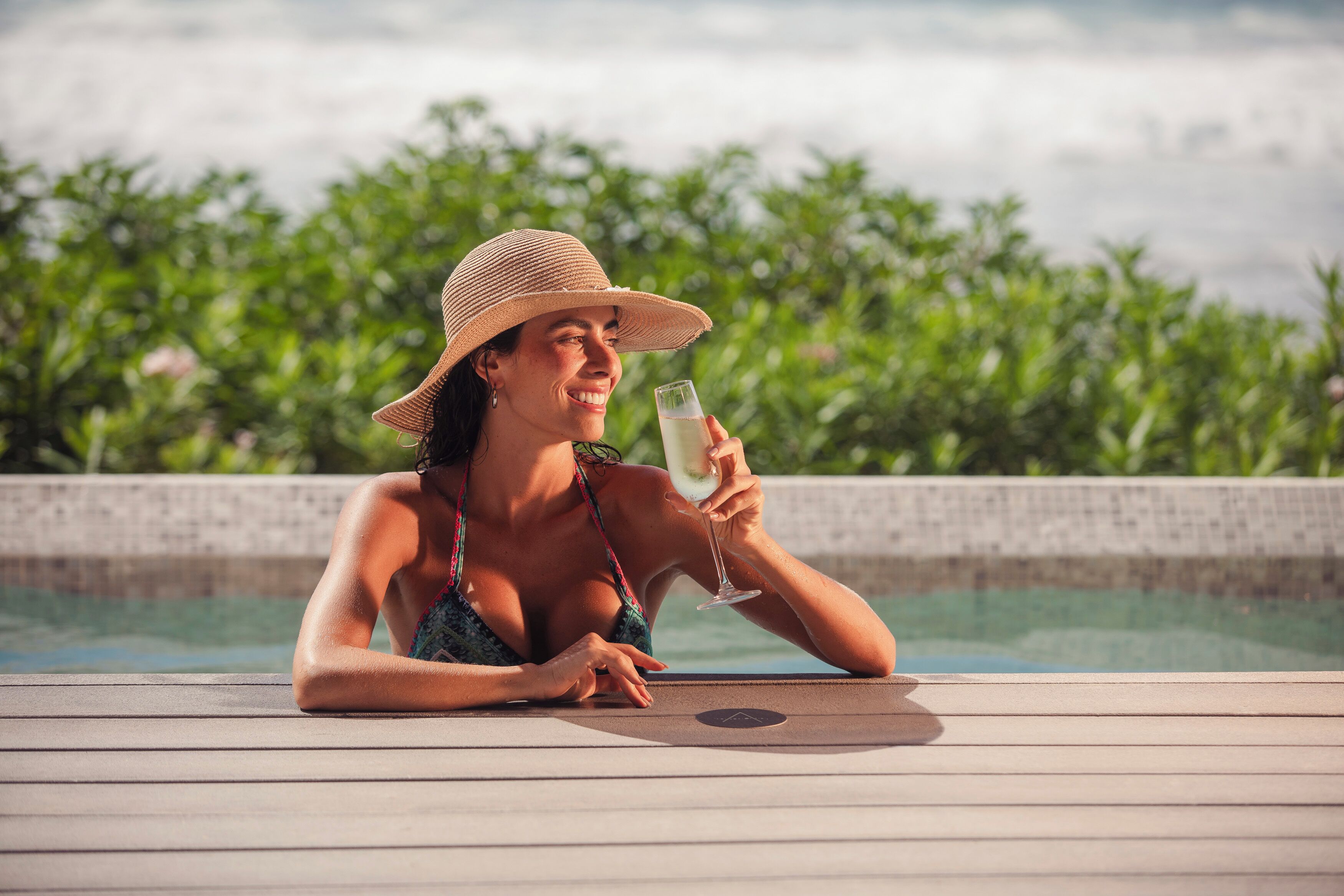 A person relaxing by a pool with a drink, surrounded by greenery.