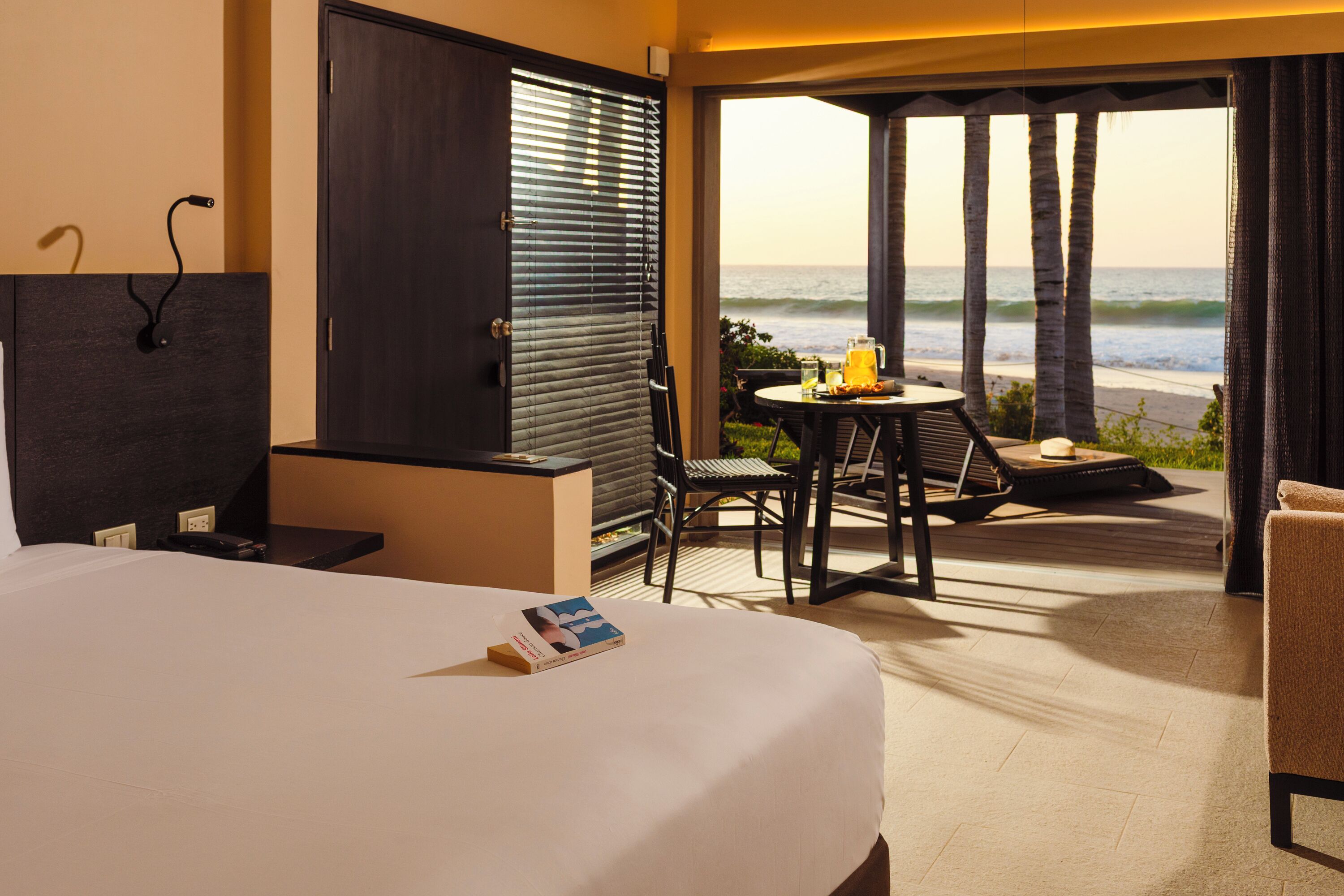 Habitación de hotel con una cama, una mesita con una bebida, una silla y vistas a la playa y al océano a través de un ventanal corredera.