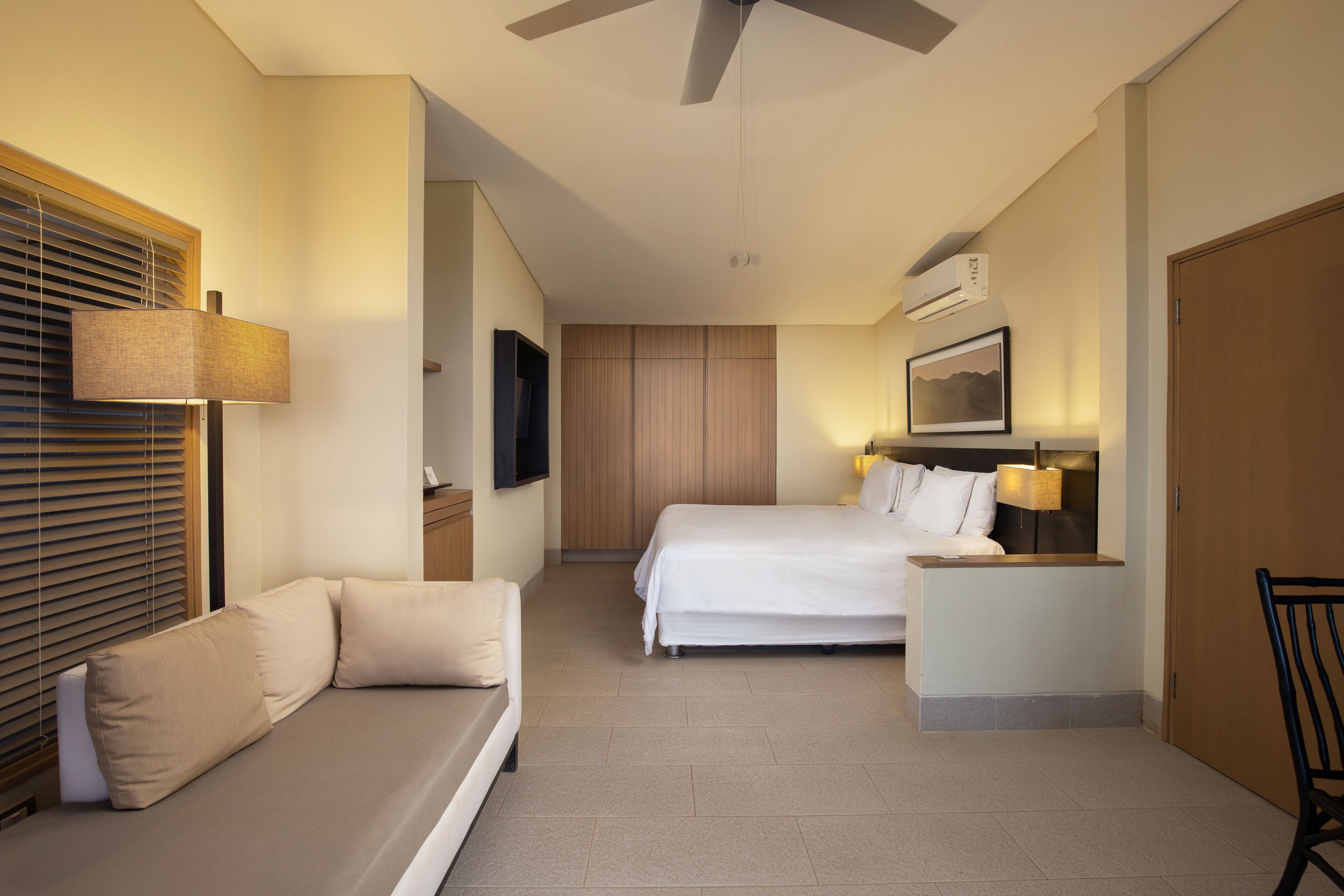 Una habitación de hotel con cama, sofá, escritorio con lámpara, televisor y ventilador de techo.