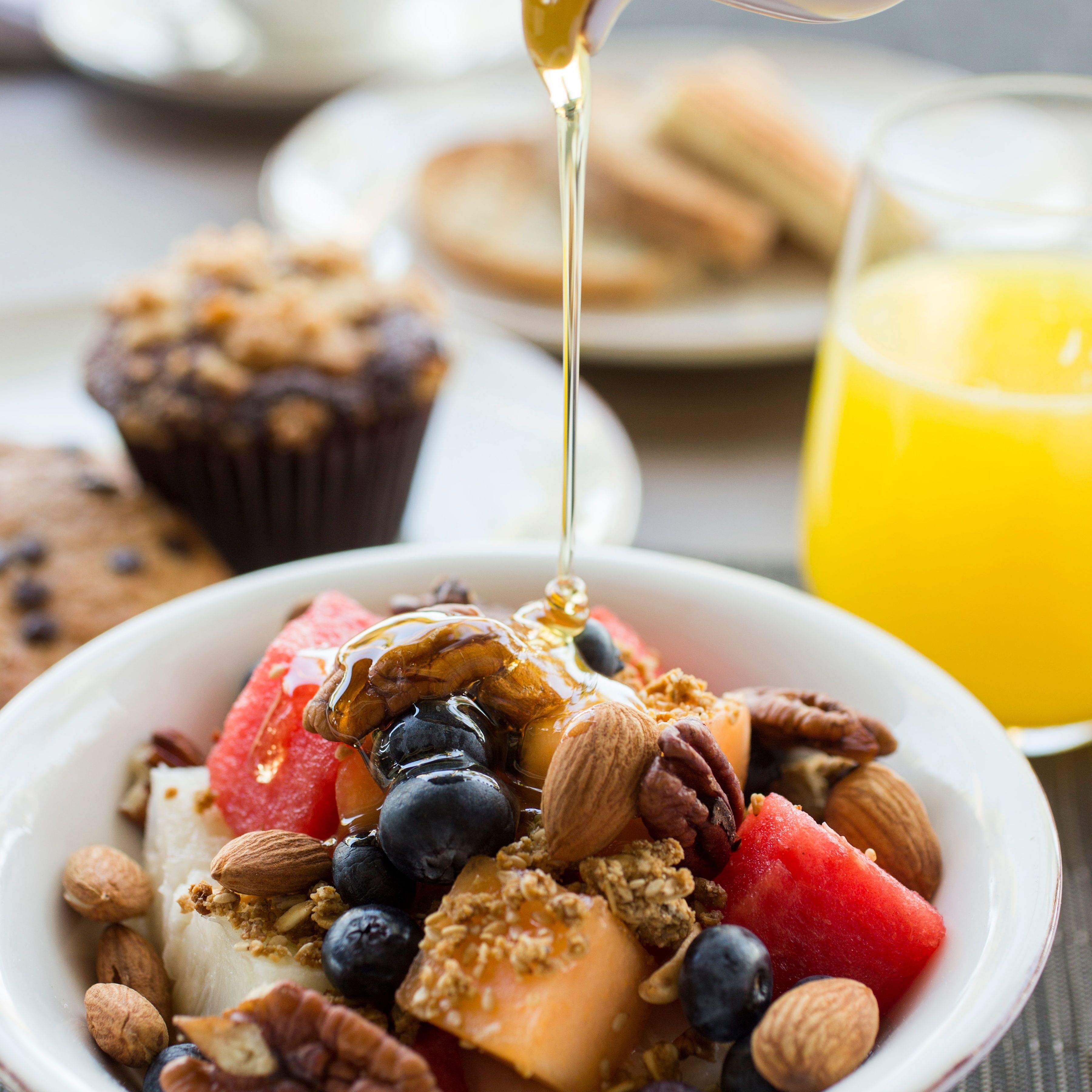 A bowl of breakfast with fruits, nuts, and granola, with syrup being poured over it.