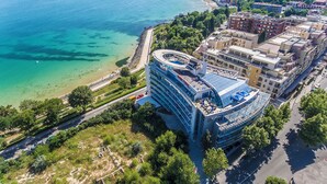 Vue aérienne d’un hôtel situé sur la côte, avec un bâtiment aux lignes courbes, une promenade en bord de mer et une vue dégagée sur l’océan.