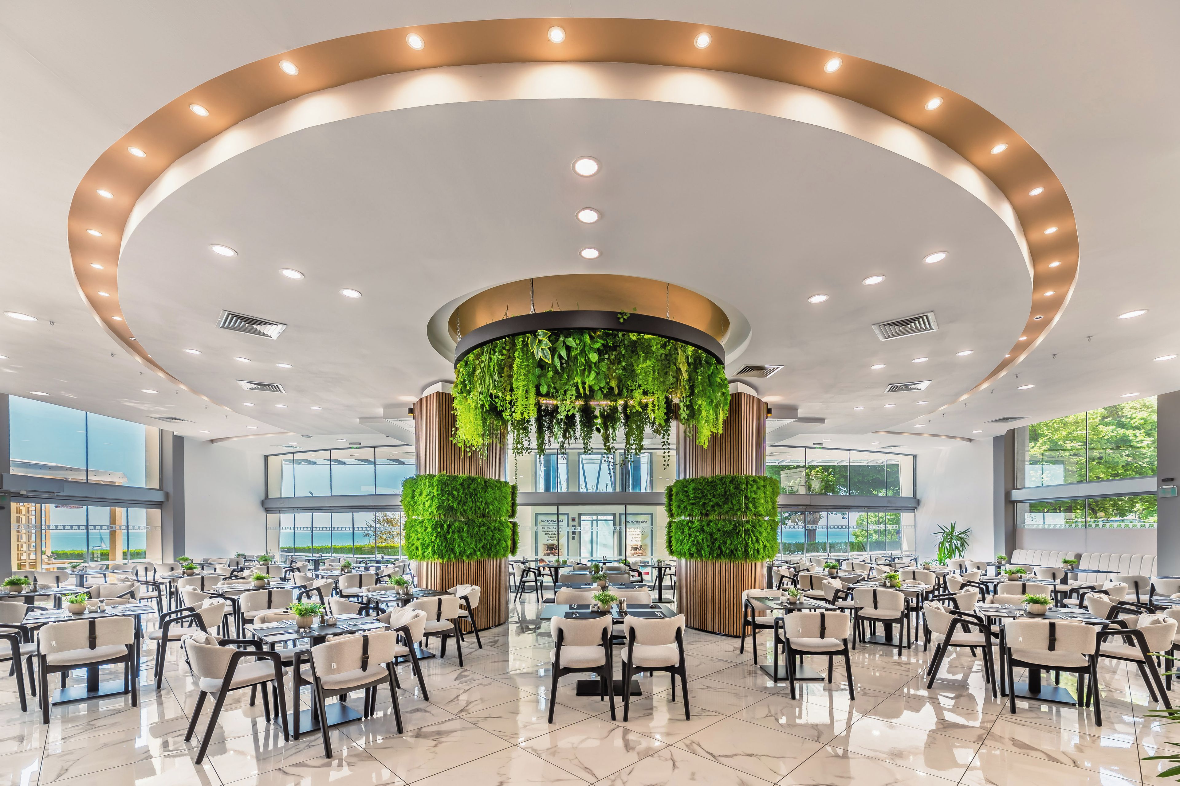 A spacious indoor dining area with multiple tables and chairs, a central column with greenery, and large windows providing natural light.