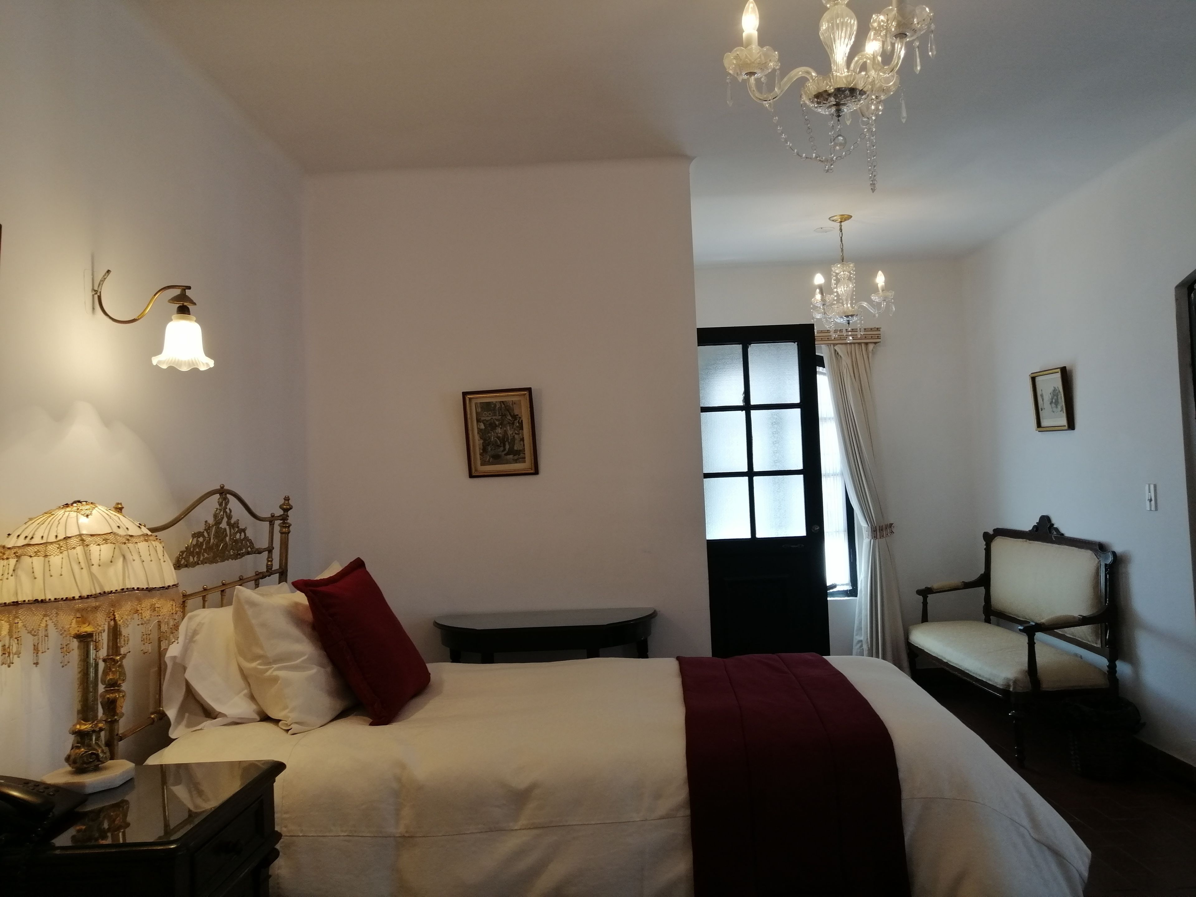 A hotel room with a bed, a desk, a chair, a chandelier, and a window with curtains.