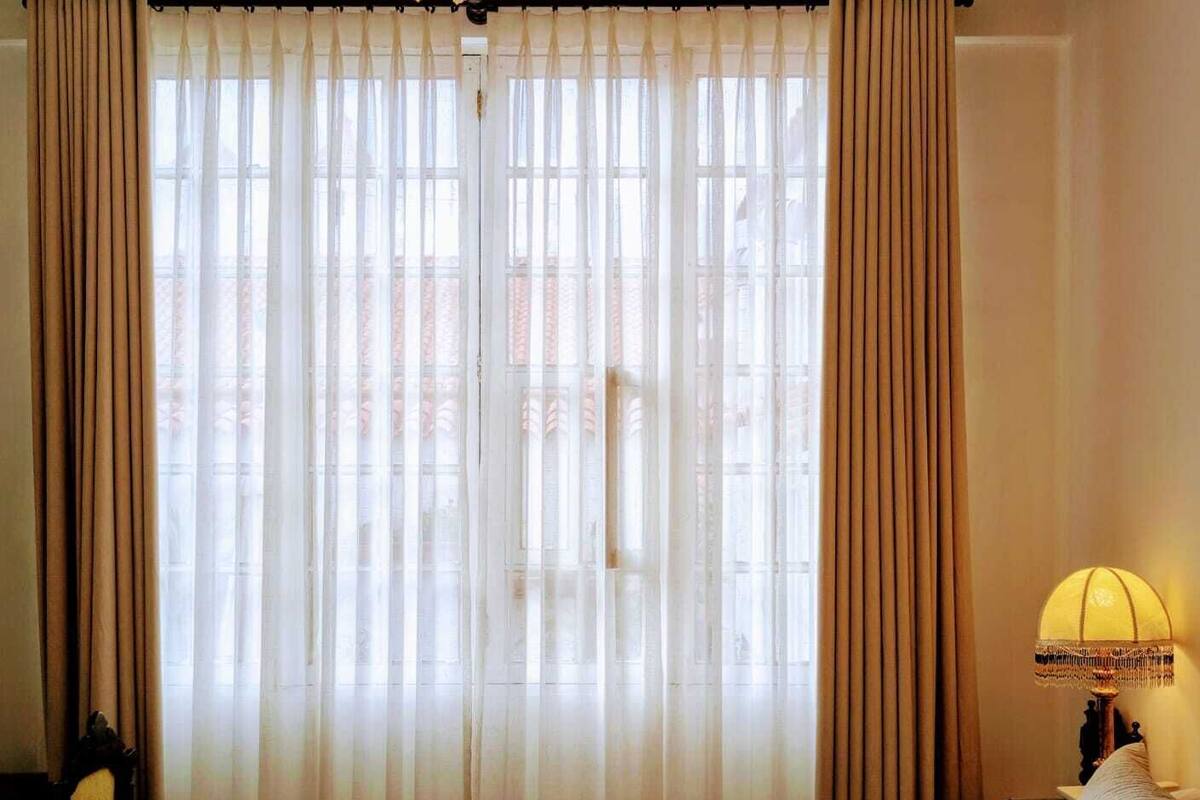 Quarto de hotel com cama, cortinas, luminária e vista pela janela.