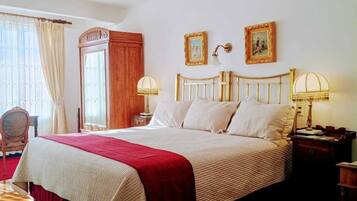 A bedroom with a large bed, wooden floor, chandelier, two wall-mounted lamps, and a door.