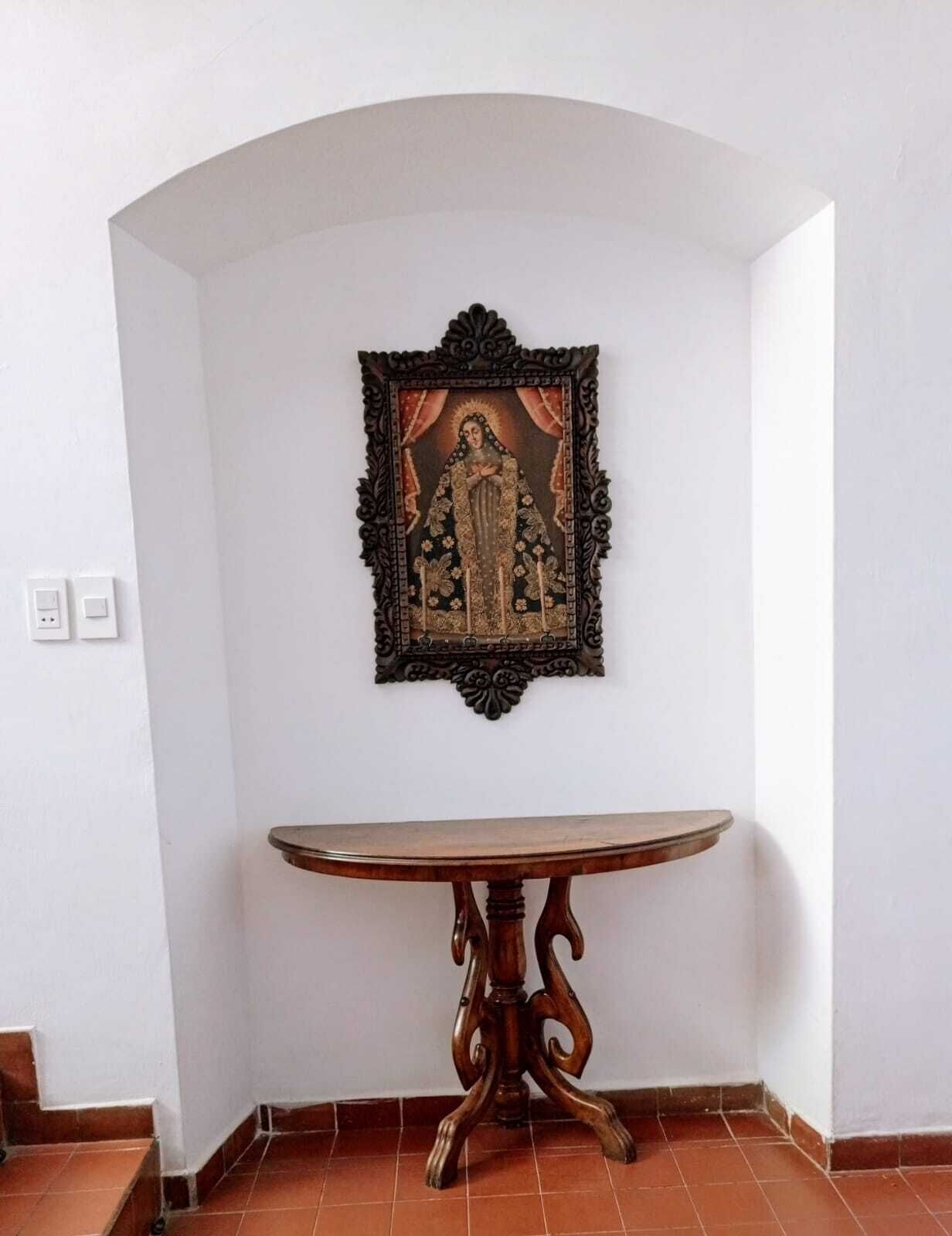 A corner room with a white wall, a framed religious painting, a wooden table, and a tiled floor.