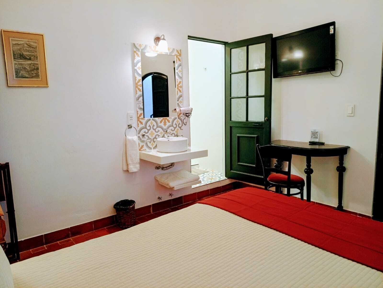A hotel room with a bed, a small table, a chair, a sink, a mirror, a television, and a framed picture on the wall.