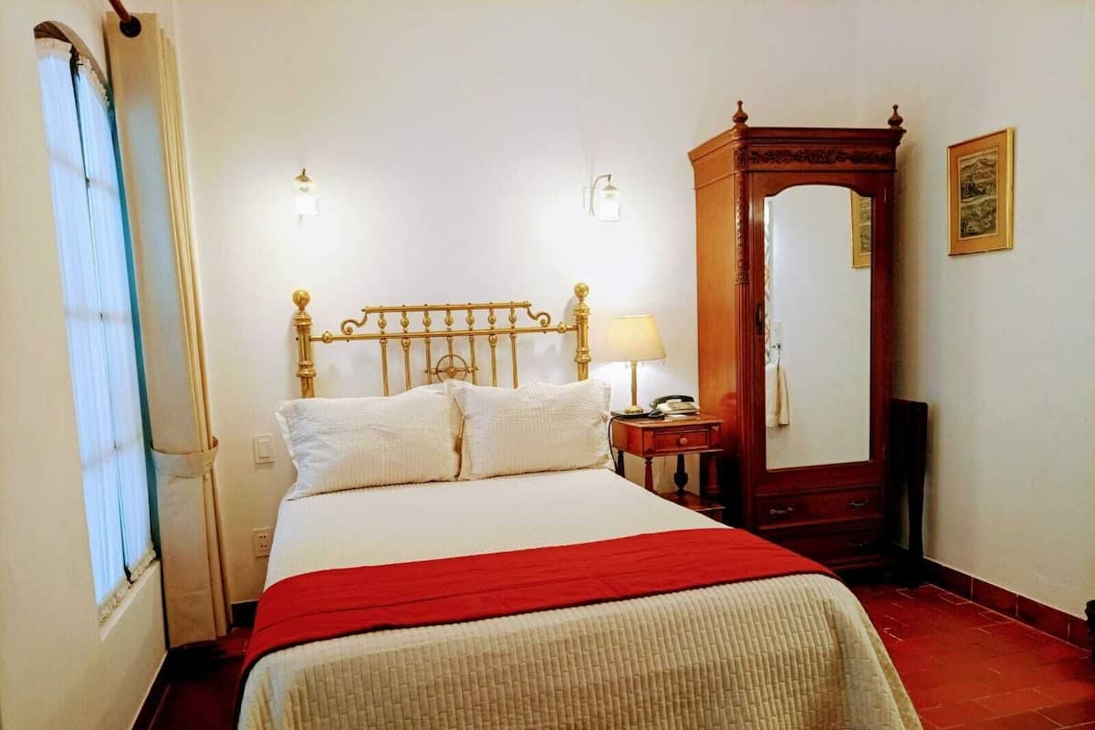 Quarto de hotel com cama, mesa de cabeceira, espelho e janela com cortinas.