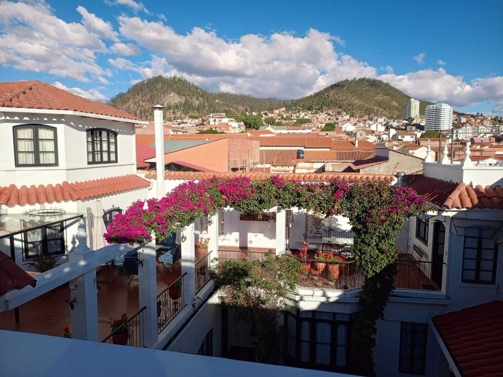 Un quartier résidentiel aux toits de tuiles rouges, avec un balcon orné de fleurs roses et une vue sur une colline.
