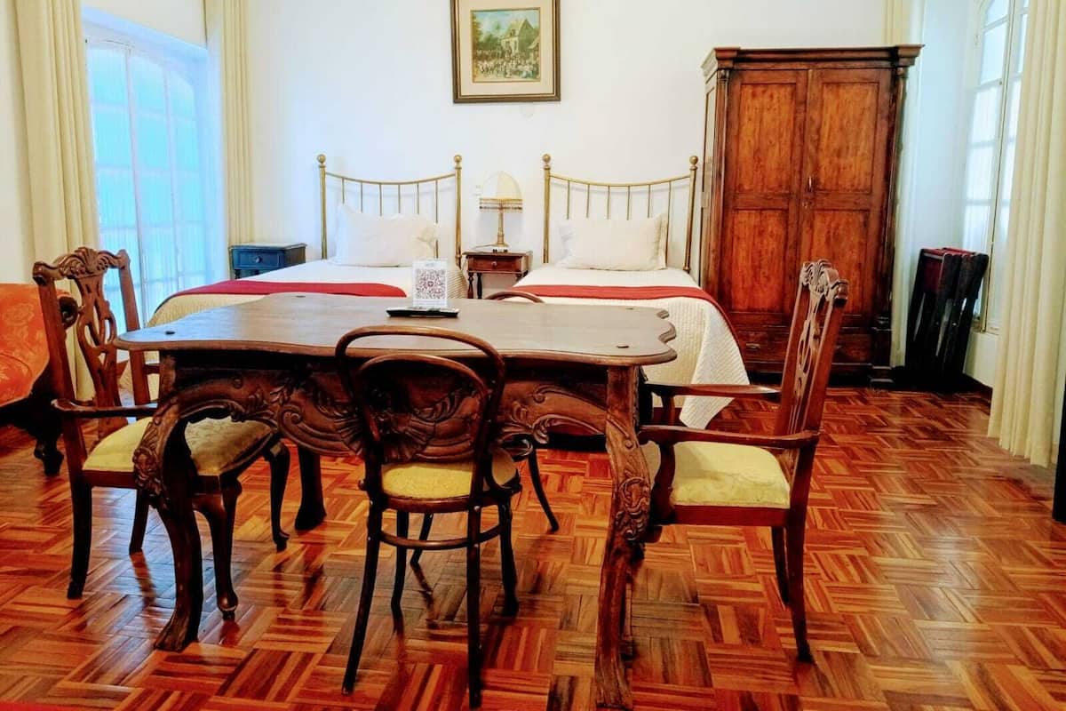 Quarto de hotel com uma cama grande, uma mesa de madeira com cadeiras, um guarda-roupa de madeira e um quadro emoldurado na parede.