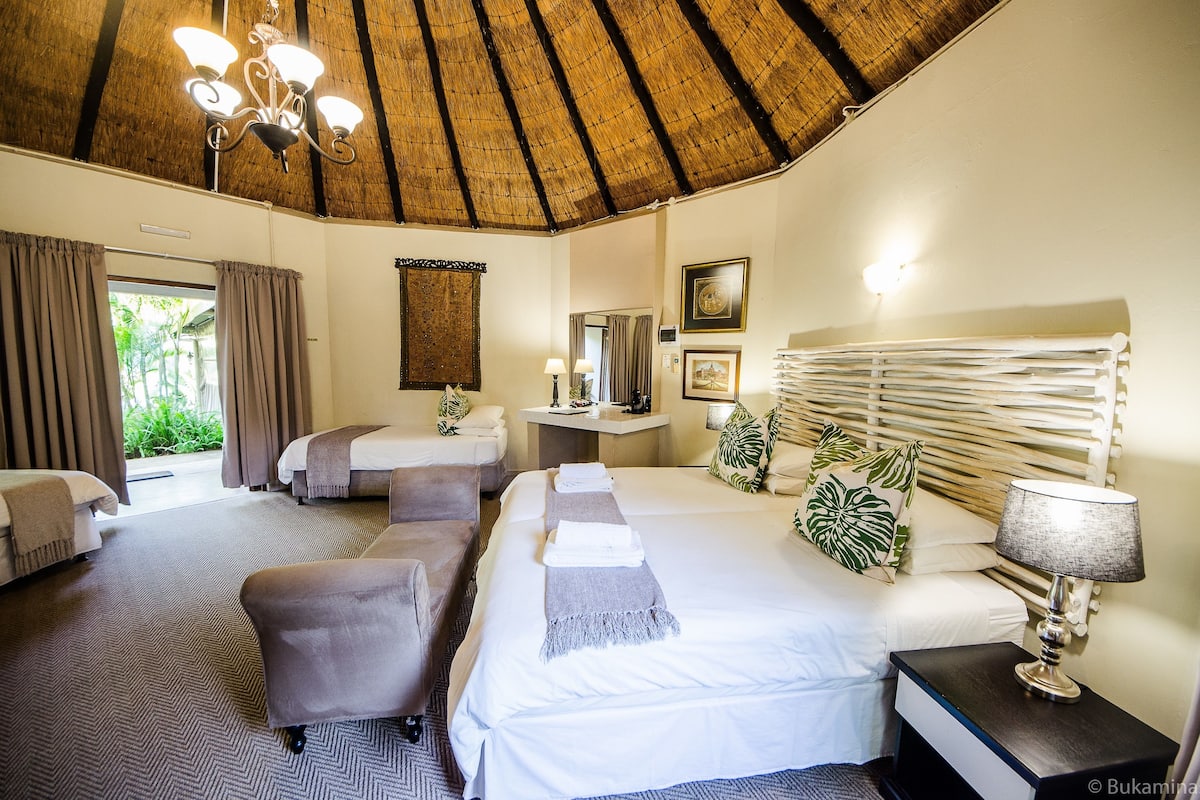 A hotel room with a thatched roof, two beds, a sitting area, and a view of greenery outside.