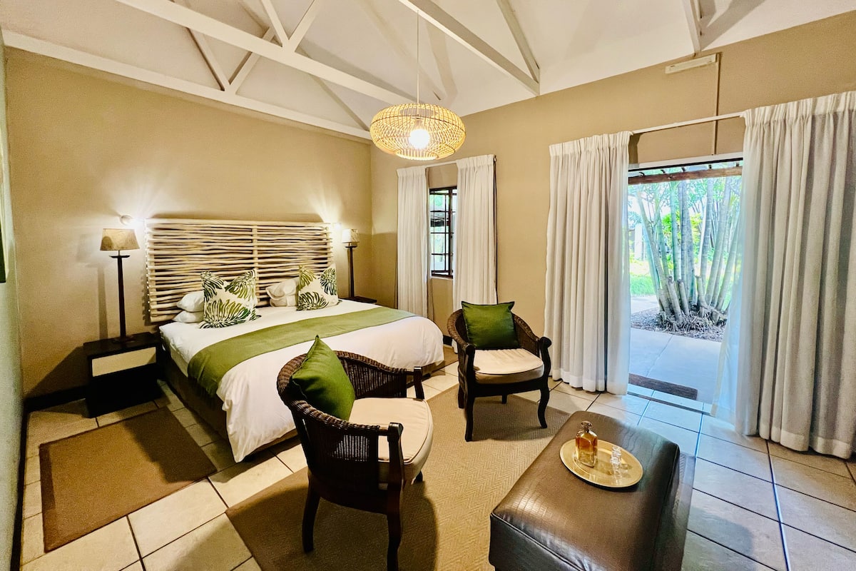 A hotel room with a bed, two chairs, a bench, a table with a tray, and a view of greenery outside.