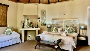 A hotel room with a thatched roof, a bed, a desk with a lamp, a chair, and a bedside table with a lamp. - Lidiko Lodge (St. Lucia)