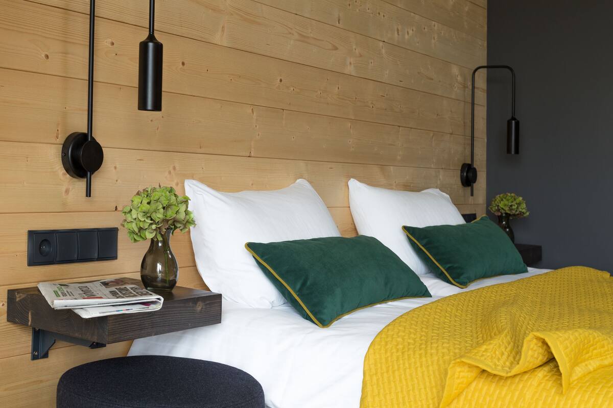 Un dormitorio moderno con cabecera de madera, una mesita de noche con un periódico, un florero con flores y una manta amarilla sobre la cama.