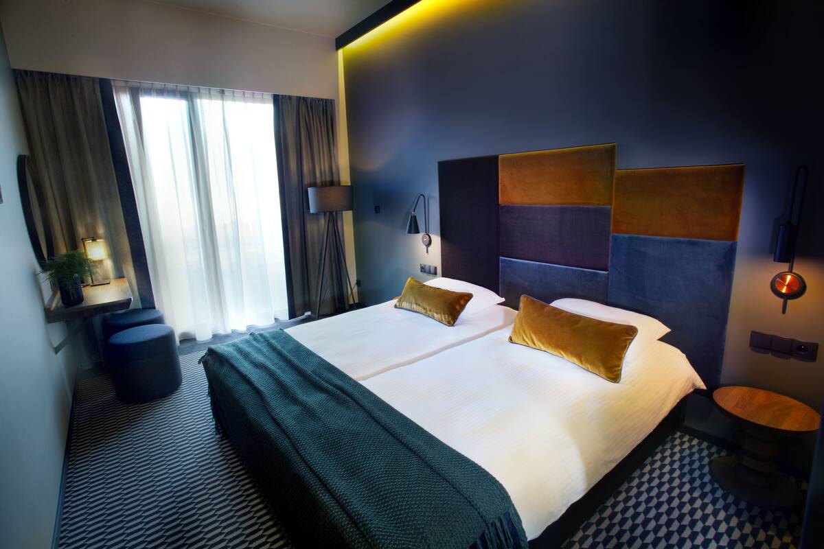 Un hotel moderno con una cama grande, dos mesitas de noche, una pequeña mesa redonda, una lámpara y vistas al exterior a través de la ventana.