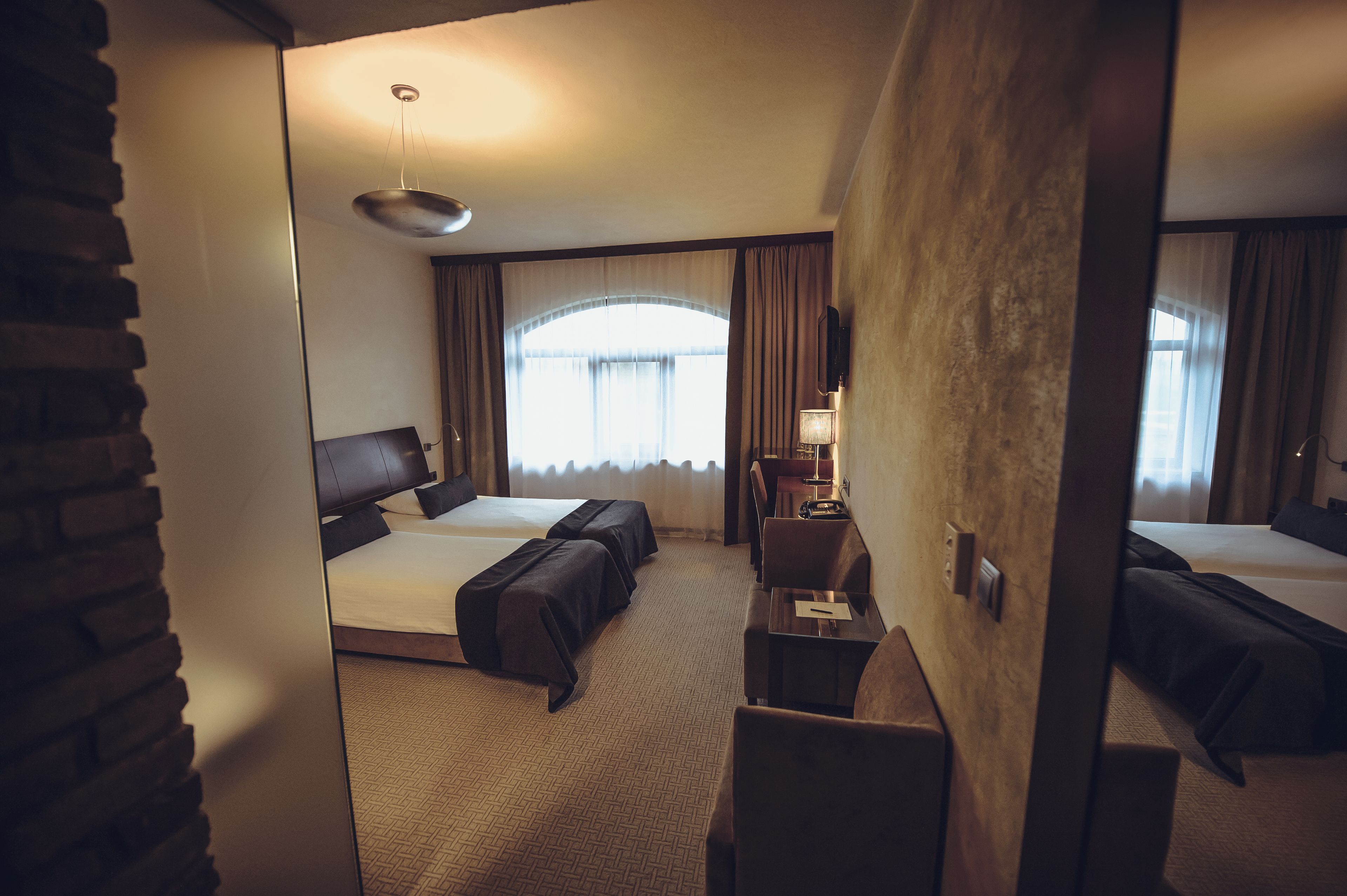 Een hotelkamer met twee bedden, een bureau, een stoel, een lamp en een groot raam met gordijnen.