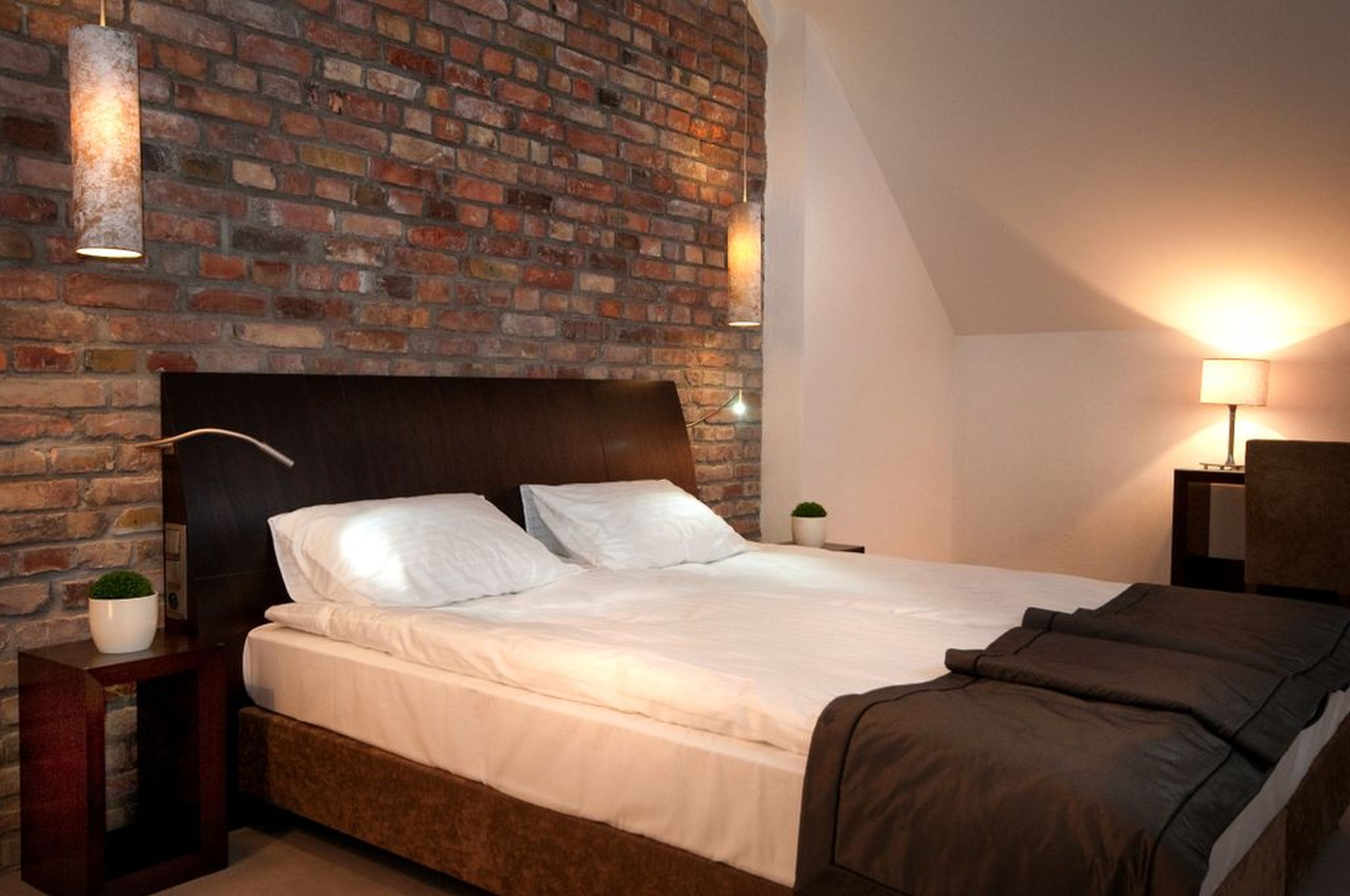 Ein Schlafzimmer mit Backsteinwand, Holz-Kopfteil, einem Bett mit weißen Leinen, einer braunen Tagesdecke und einem Nachttisch mit Pflanze.