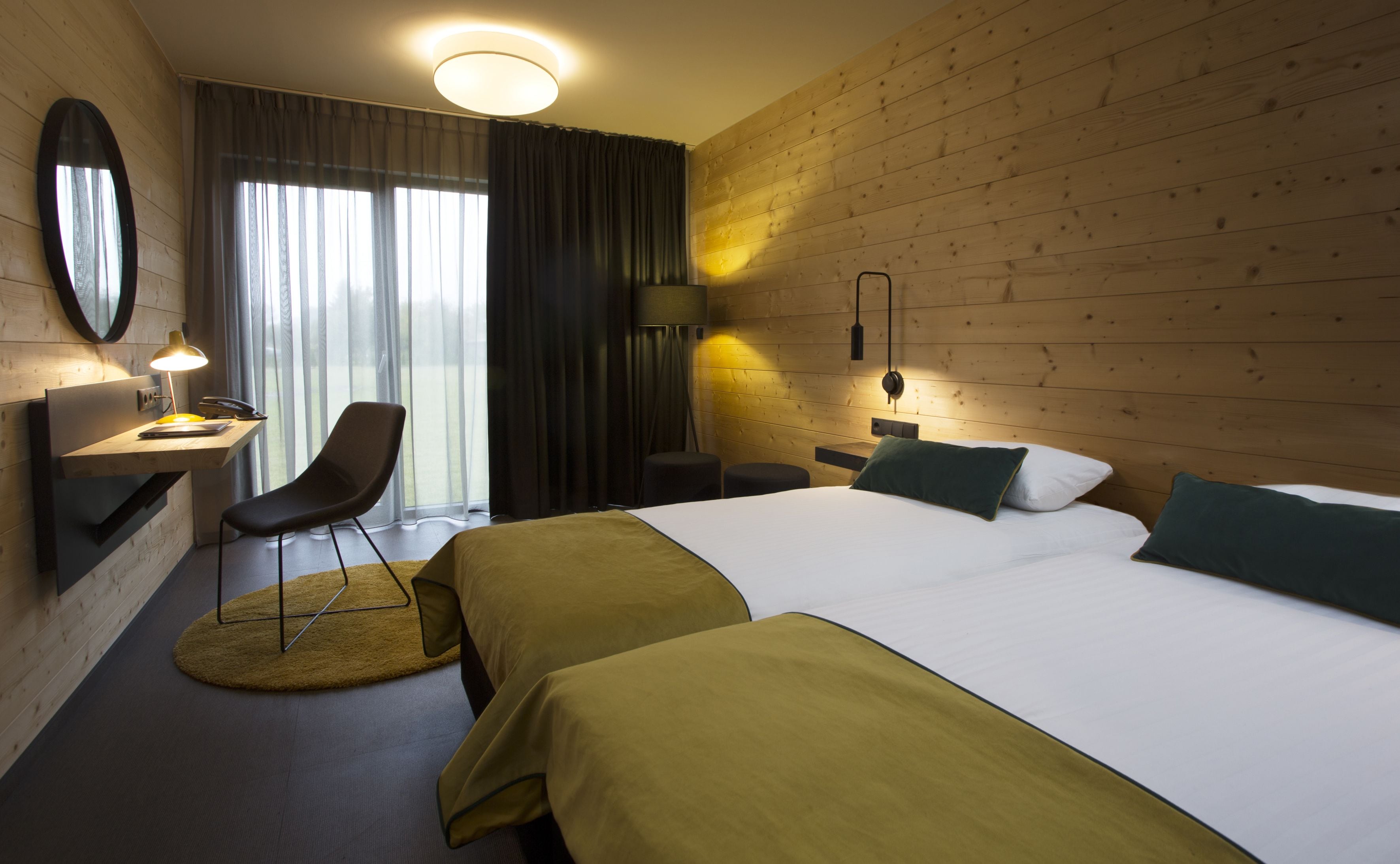 A hotel room with a large bed, two bedside tables, a desk with a lamp, a chair, and a large window with curtains.