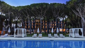 Uma área à beira da piscina com espreguiçadeiras e guarda-sóis, uma piscina e um edifício com várias varandas ao fundo.