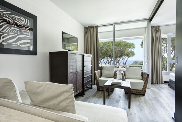 A modern living room with a sofa, armchairs, a coffee table, and a cabinet. Large windows offer a view of trees and the sea.