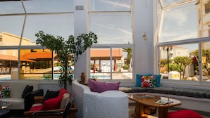 Un espacio interior con mesas y sillas iluminado por el sol, con vistas a una piscina y edificios.